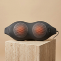ArchZen heated lumbar massage cushion, slim dark-gray peanut shape with two dimpled massage zones, on a travertine pedestal