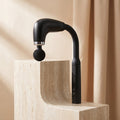 Black PulseZen curved-handle massage gun on a travertine pedestal